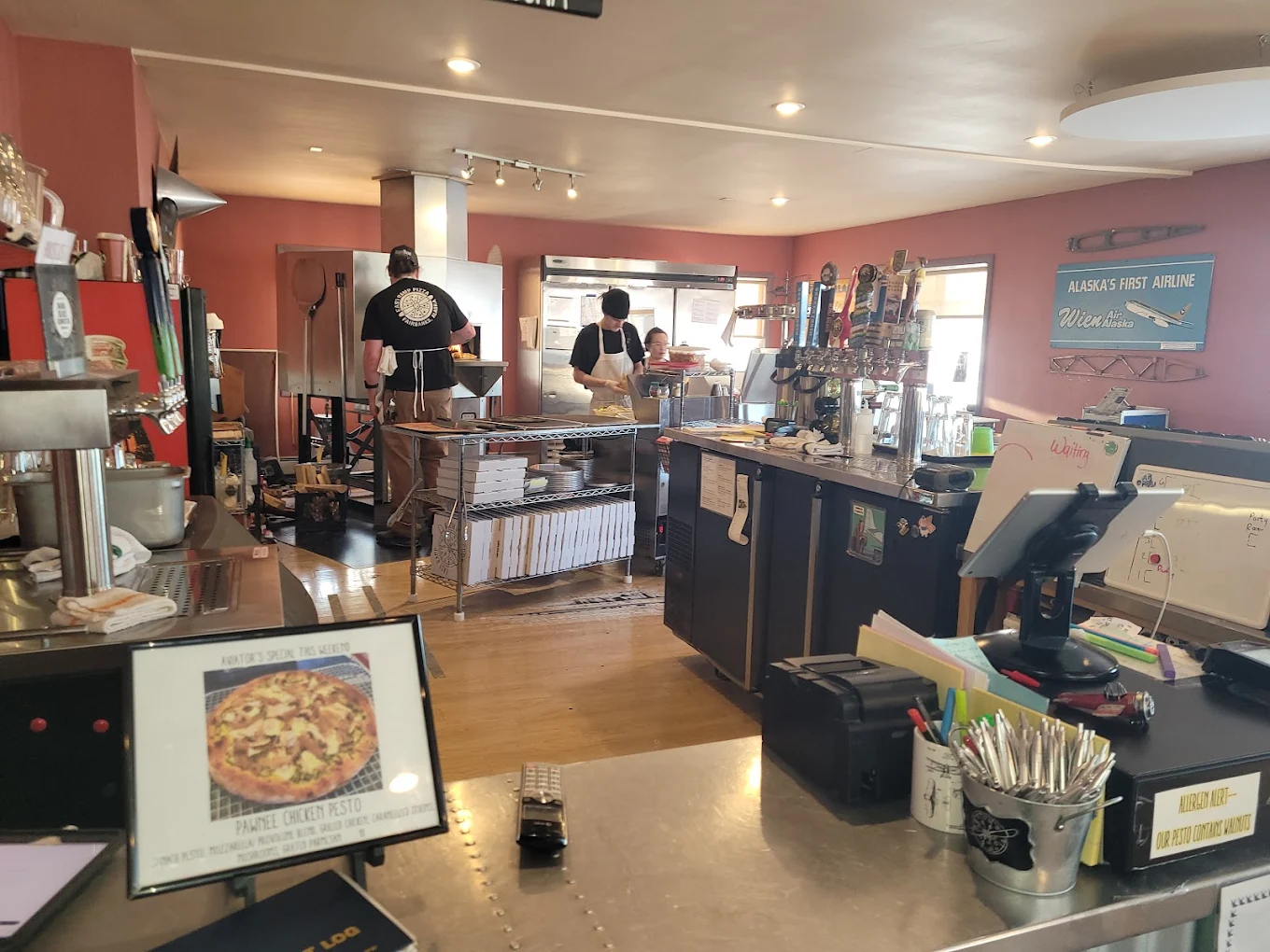 Inside East Ramp — pizzaiolos working the line in front of the Woodstone oven, beer taps on the right, dusty-rose walls and a Wien Air Alaska sign on the wall
